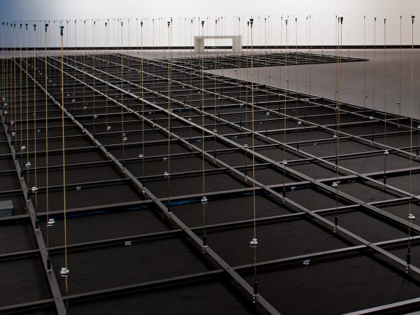 Gallery installation of black panels on the floor with a grid of gray with then brass rods with small speakers mounted on top.