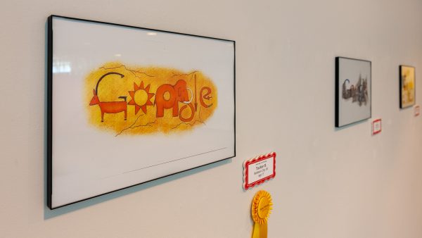 Gallery installation of a framed Google doodle orange animal figures on a yellow background. A gold ribbon is displayed to the lower right of the framed artwork.