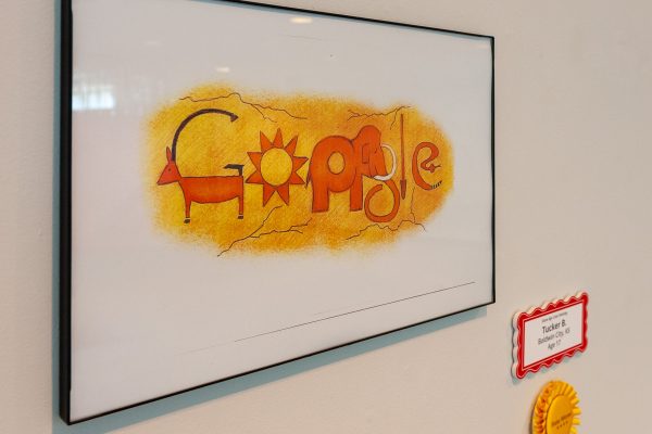 Gallery installation of a framed Google doodle orange animal figures on a yellow background. A gold ribbon is displayed to the lower right of the framed artwork.