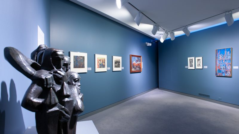Gallery installation with a black sculpture of people in the foregound and a other framed artwork ona blue wall.