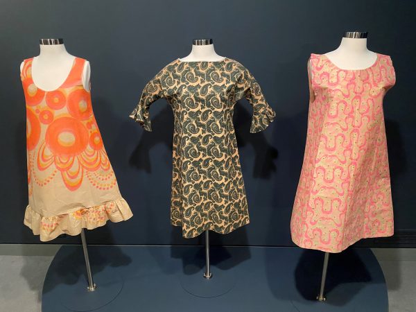 Three paper dresses on display stands, the left one sleeveless with orange circles, the center one long-sleeved with a grey print patter and the far right one short-sleeved with pink and ivory designs