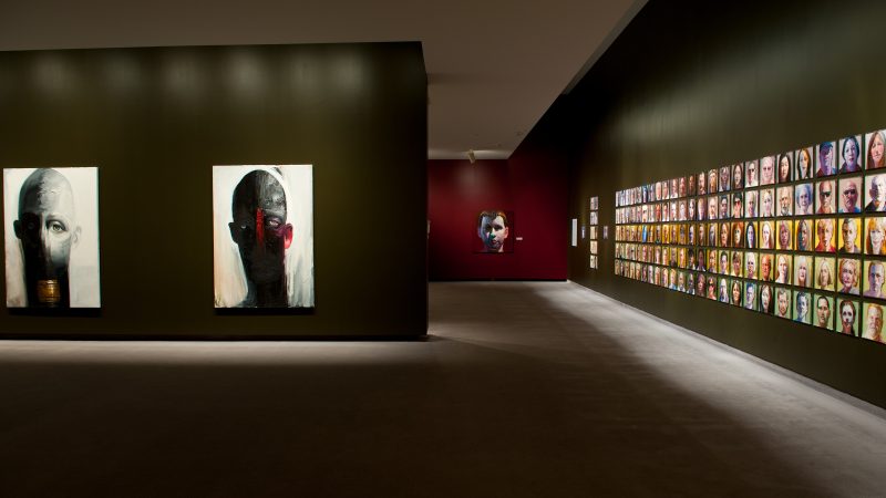 Gallery image with a wall with large scale portraits on the left and a long wall of smaller portraits on the right