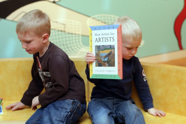 Two little boys who are white with blonde hair sit on a wooden bench facing away from each other while one of them holds up a book. They have on dark long-sleeve shirts and jeans.