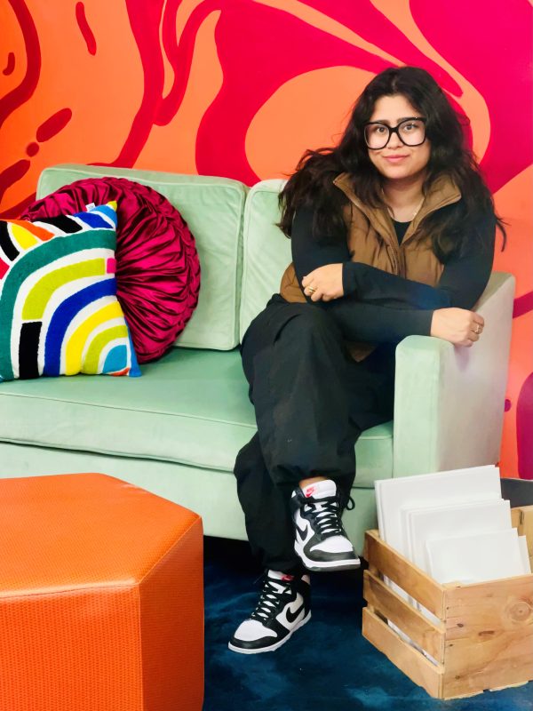 woman sitting on a mint green couch against a bright two-toned patterned orange wall with an orange ottoman in front. She has long dark hair, wears glasses, and is dressed in black and wearing black and white tennis shoes