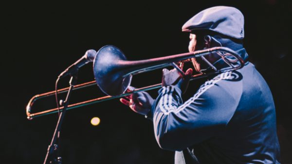Photo of a Black man with his back towards the camera while facing the audience and playing trombone in front of a mic