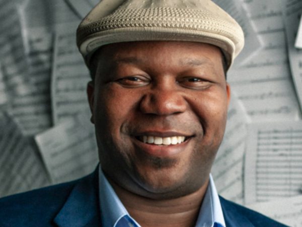 Photo of a Black man smiling and looking at the camera wearing a light brown hat, light blue shirt and dark blue jacket