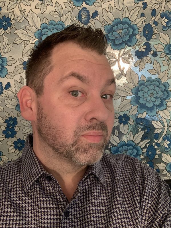 Photo of a white man with short brown hair looking at the camera wearing a dark grey shirt in front of blue and silver floral wallpaper