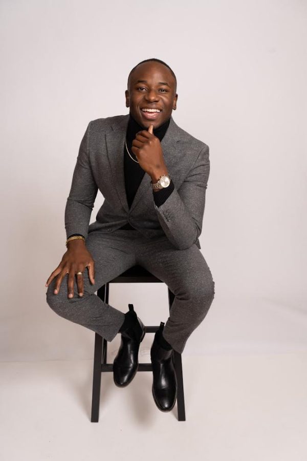 Photo of a Black man sitting on a stool, smiling for the camera and wearing a dark grey suit with a black shirt, black shoes and a silver watch
