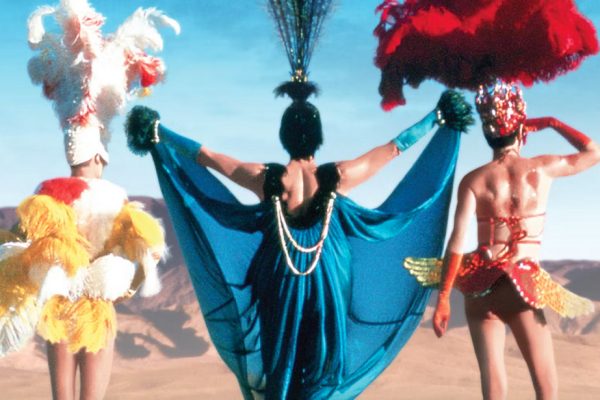 Three men in colorful costumes of yellow, blue and red stand together with their backs toward the camera and look out over the desert