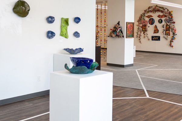 Gallery image with blue sculpture on a white pedastal in the foreground, various other artworks on the walls