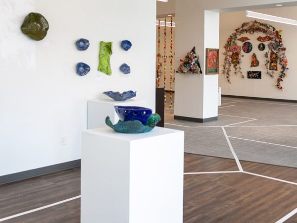 Gallery image with blue sculpture on a white pedastal in the foreground, various other artworks on the walls