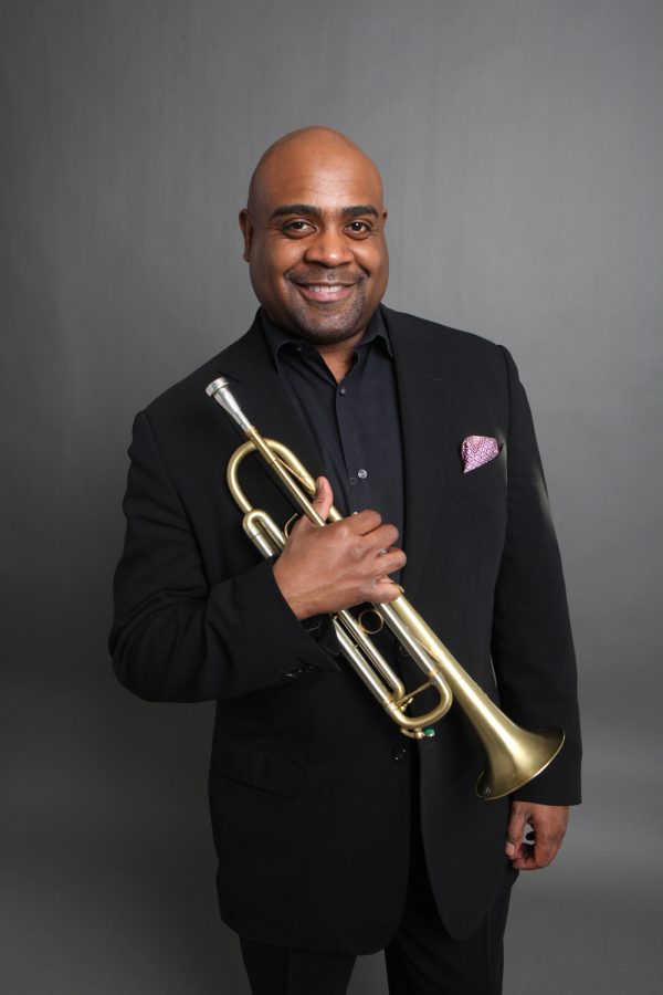 Photo of a Black man wearing a black suit smiling for the camera and holding a trumpet
