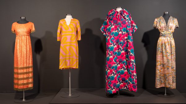 Installation image of four colorful dresses hanging on display forms: the first short-sleeve dress is long, with orange and gold; the second long-sleeve dress is short with an orange and pink pattern; the third short-sleeve jacket dress has a purple, teal, white and pink design; the fourth short-sleeve, v-neck dress has a shimmery pattern in pastel colors