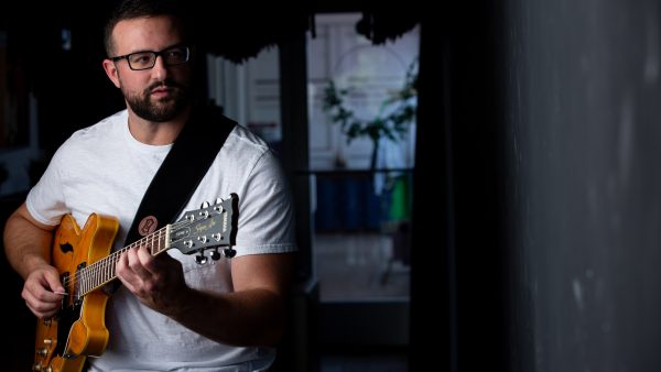 Man with dark framed glasses and a close trimmed beard, dressed in a white t-shirt, plays a guitar. Standing in front of a dark background with a reflection of a window