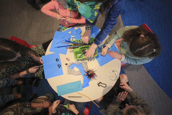 Several children sit around a round table with blue sheets of paper and markers