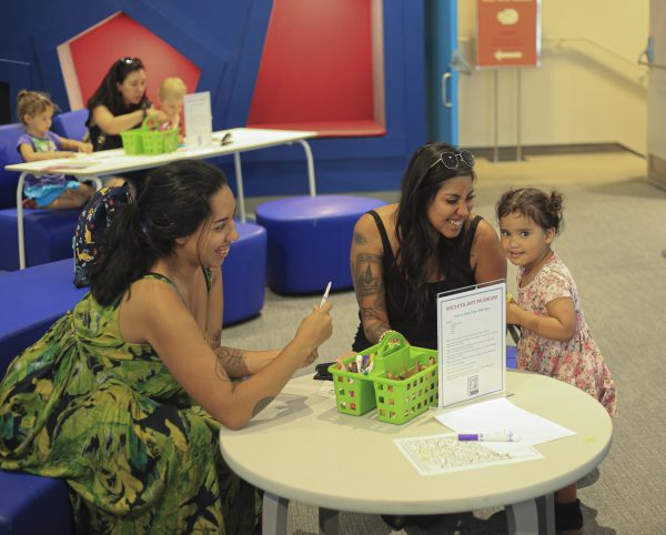 Two women and a little girl sit around a table in the Living Room and work on coloring sheets with a mother and two young children in the background