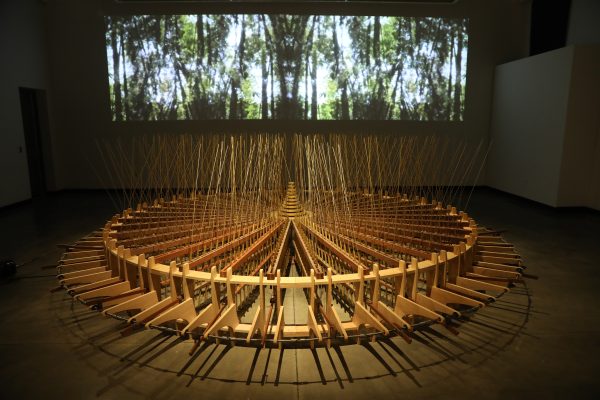 Art installation on the floor with a large, round wooden rotary shape and a video projection of a forest in the background