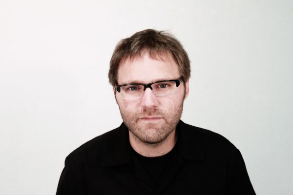 Photo of a man against a white background looking at the camera with brown hair glasses and a dark shirt