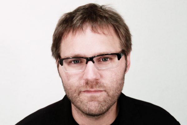 Photo of a man against a white background looking at the camera with brown hair glasses and a dark shirt