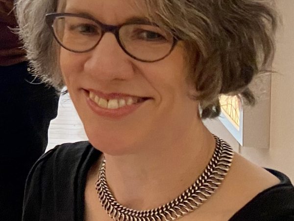 Woman with chin-lengthy light grey hair wearing glasses, a necklace and a black top