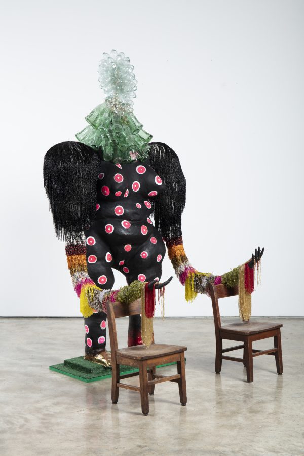 Photo of the sculpture by vanessa german showing a figure and hands outstretched and balanced on two small wooden chairs