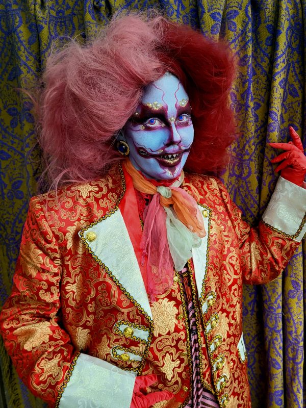 Person dressed as a clown with puffy orange hair, white face and dressed in an orange print jacket with wide white lapels.