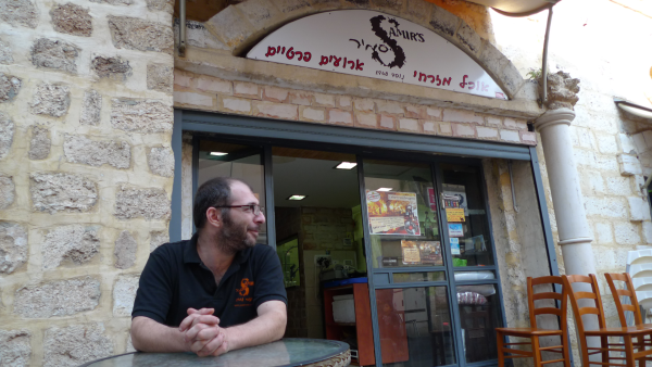 Photo for Hummus the Movie. The Host is seated next to a stone wall outside a Jewish restaurant.