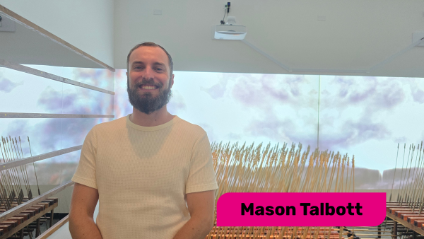 alt= Mason Talbott posing in front of a mechanical wheat exhibition at WAM