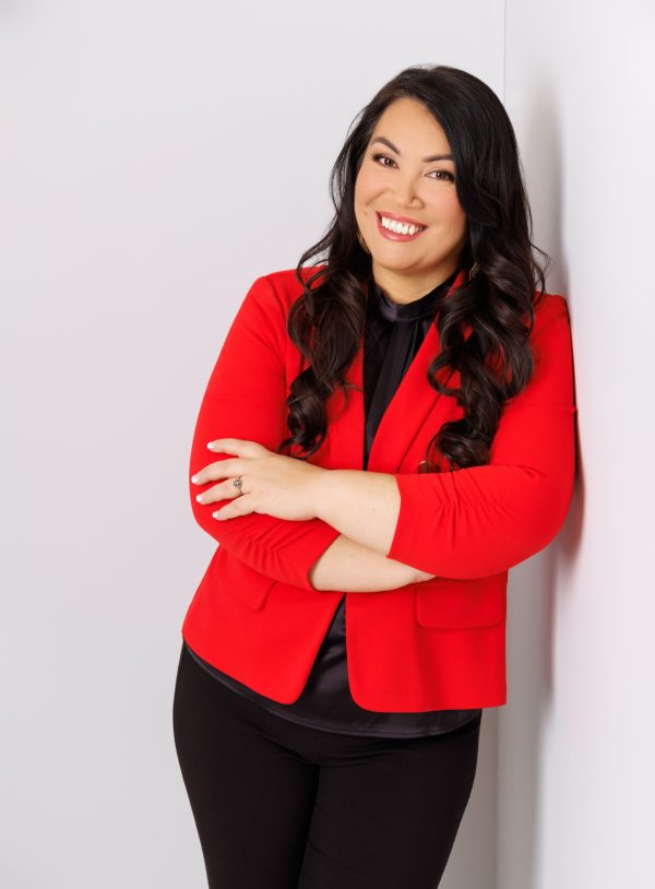 alt= headshot of Yeni Silva-Renteria, wearing a bright red jacket, black pants, arms crossed, leaned against white wall, smiling at camera